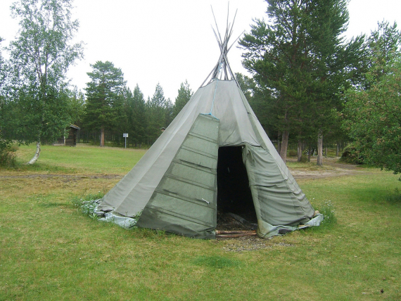 Fotografi av en oppsatt tradisjonell lavvo med åpen dør, på en gresslette i skogen.